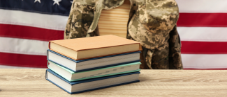 stack of books on desk in front of empty chair on which hangs a camaflauge jacket with an American flag background